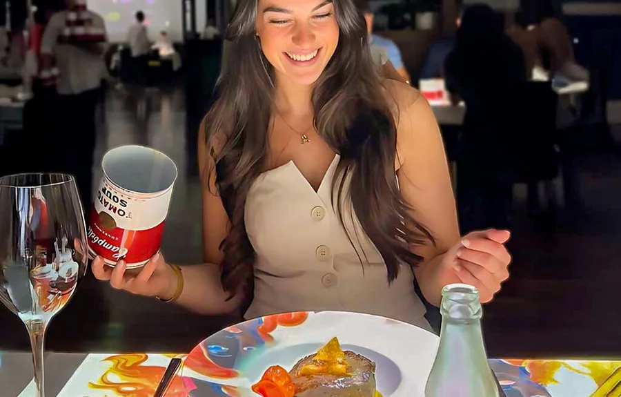 Una visitante sostiene la lata de sopa de tomate Campbell’s del plato inspirado en Warhol en una mesa iluminada por proyecciones